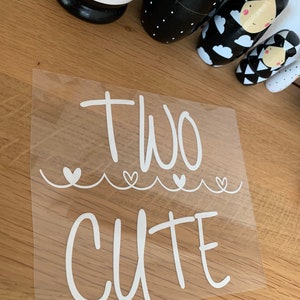 Könnte beinhalten: Weißer Vinyl-Aufkleber mit dem Text "TWO CUTE" in einer handschriftlichen Schriftart. Der Text ist von drei kleinen Herzen umgeben.