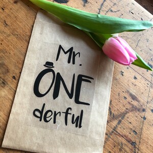 Könnte beinhalten: Eine braune Papiertüte mit einem Aufkleber, auf dem "Mr. ONE derful" in schwarzer Schrift steht. Eine rosa Tulpe liegt auf der Tüte.