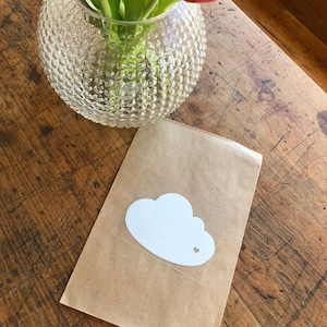 Op de afbeelding: Een bruine papieren zak met een witte sticker in de vorm van een wolk met een klein uitgesneden hartje. De zak ligt op een houten oppervlak.