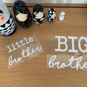 Könnte beinhalten: Zwei weiße Vinyl-Aufkleber mit den Worten "kleiner Bruder" und "GROSSER Bruder" auf einer Holzoberfläche. Die Aufkleber sind in einer kursivschrift.