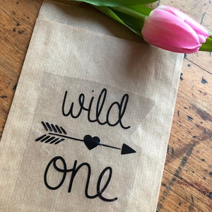Könnte beinhalten: Eine braune Papiertüte mit einem durchsichtigen Aufkleber, auf dem in schwarzer Kursivschrift "wild one" steht. Der Aufkleber zeigt außerdem einen schwarzen Pfeil mit einem Herz in der Mitte.