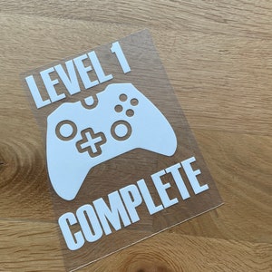 Könnte beinhalten: Weißer Vinyl-Aufkleber mit einem Videospiel-Controller und dem Text "LEVEL 1 COMPLETE". Der Aufkleber befindet sich auf einer transparenten Rückseite, die auf einer hellbraunen Holzoberfläche liegt. Für Gamer.