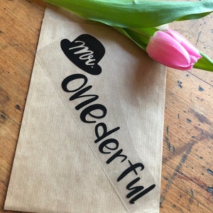 Könnte beinhalten: Ein schwarz-weißer Aufkleber mit dem Text "moi." und "wonderful" in einer kursivschrift. Der Aufkleber befindet sich auf einem braunen Papierbeutel mit einer rosa Tulpe.