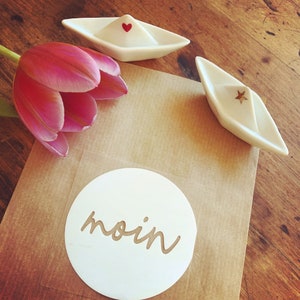 Könnte beinhalten: Ein weißer Papierkreis mit dem Wort "moin" in Kursivschrift darauf. Der Kreis liegt auf einer braunen Papieroberfläche. Es gibt zwei kleine weiße Keramikboote mit einem roten Herzen und einem goldenen Stern darauf.