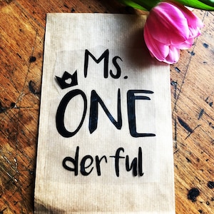 Könnte beinhalten: Eine braune Papiertüte mit dem schwarzen Schriftzug "Ms. ONE derful" und einer kleinen Kronengrafik. Eine rosa Tulpe liegt oben rechts. Das Ganze liegt auf einer Holzoberfläche.