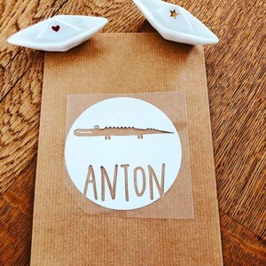 Op de afbeelding: Een witte ronde sticker met een bruine alligator en de naam Anton in wit gedrukt.