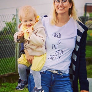 Op de afbeelding: Een vrouw draagt een wit T-shirt met de tekst "Tired is the new pretty" en houdt een peuter vast die een beige jas en een gele broek draagt. De vrouw draagt een blauwe jeans, een gestreepte cardigan en een bril.