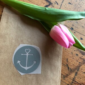 Könnte beinhalten: Eine hellbraune Papiertüte mit einem weißen Ankeraufkleber. Der Anker ist weiß mit einem Herz in der Mitte. Eine rosa Tulpe liegt auf der Tüte.