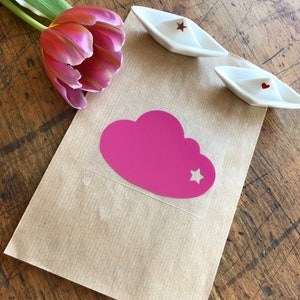 Op de afbeelding: Een bruine papieren zak met een roze wolk sticker met een witte ster uitsparing. De sticker zit op een transparante plastic vel. Er zijn twee witte papieren bootjes met rode sterren op de zak.