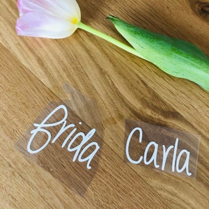 Könnte beinhalten: Zwei durchsichtige Plastik-Platzkarte mit den Namen "Frida" und "Carla" in weißer Schreibschrift. Eine rosa und weiße Tulpe mit grünen Blättern liegt auf einer Holzoberfläche hinter den Platzkarte.