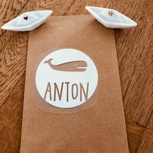 Könnte beinhalten: Ein weißer runder Aufkleber mit einer Wal-Silhouette und dem Namen Anton in Schwarz gedruckt. Der Aufkleber befindet sich auf einem braunen Papierhintergrund.