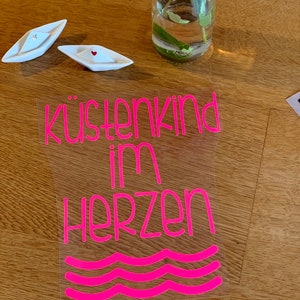 Könnte beinhalten: Pinkfarbener Vinyl-Aufkleber mit dem Text "küstenkind im herzen" und drei pinkfarbenen Wellenlinien darunter. Der Aufkleber befindet sich auf einer Holzoberfläche.