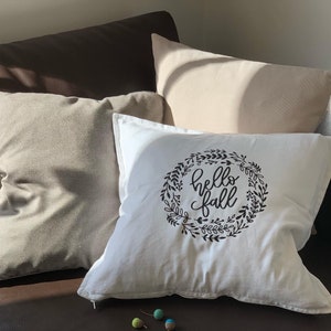 Könnte beinhalten: Ein weißes Kissen mit einem schwarzen Kranz-Design und dem Text "hello fall" in einer handschriftlichen Schrift.
