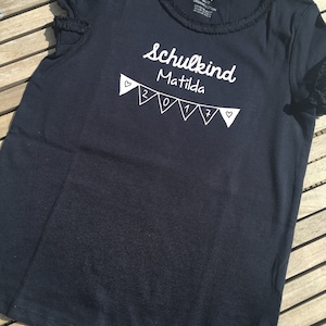 Könnte beinhalten: Schwarzes T-Shirt mit weißem Text "Schulkind Matilda 2017" und einer Girlande mit Fahnen und Herzen an den Enden.