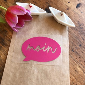 Könnte beinhalten: Eine braune Papiertüte mit einem rosa Sprechblasensticker, auf dem "moin" steht. Auf der Tüte liegen zwei weiße Papierboote mit roten Herzen und Sternen.