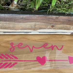 Könnte beinhalten: Rosa Vinyl-Aufkleber mit dem Wort "seven" und einem Pfeil mit einem Herz in der Mitte. Der Pfeil hat ein rosa-weiß gestreiftes Muster.
