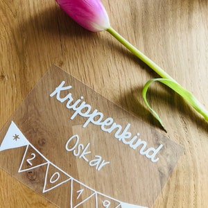 Könnte beinhalten: Ein klares Acrylschild mit weißer Beschriftung, auf dem "Krippenkind Oskar 2019" steht, mit einer Girlande aus weißen Wimpeln mit Sternen an jedem Ende.