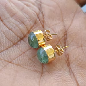 以下が含まれることがあります： ライトグリーンの丸みを帯びた宝石が付いた、金色のスタッドピアス。 イヤリングは金のベゼルセッティングで、ポストとバタフライクラスプが付いています。 イヤリングは、明るい茶色の表面に表示されています。