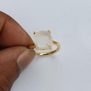 Puede incluir: Un anillo de oro con una gema translúcida grande, de corte cuadrado. La banda y el engaste del anillo son de color oro brillante, creando un contraste llamativo con la piedra pálida. La gema está sujeta por cuatro garras.