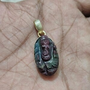 May include: A gold pendant with a carved gemstone featuring a multi-colored design. The gemstone is oval-shaped and has a dark green, blue, and red color scheme.