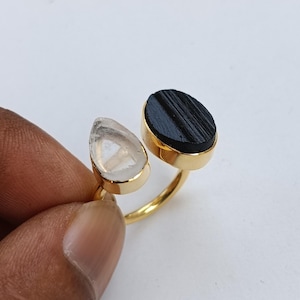 以下が含まれることがあります： 2つの宝石をあしらったゴールドカラーのリング。1つは透明な涙の形をした宝石、もう1つは黒い楕円形の石です。リングのバンドは細く金色で、宝石は金色のベゼルにセットされています。