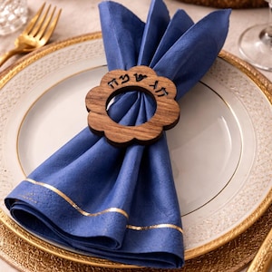 May include: A blue linen napkin with gold trim, held by a wooden flower-shaped napkin ring with Hebrew text. The napkin rests on a white plate with a gold rim, atop a gold charger. A gold fork is visible.