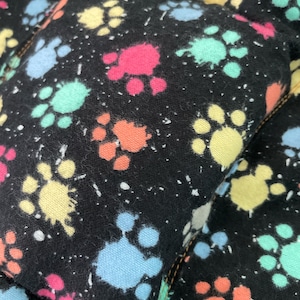 May include: Close-up of a black fabric with colorful paw prints in pink, yellow, blue, orange, and green. White speckles are scattered across the fabric. The fabric is stitched with a brown thread.
