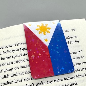 Könnte beinhalten: Ein Lesezeichen mit der Flagge der Philippinen. Das Design zeigt ein weißes Dreieck mit einer gelben Sonne und drei Sternen sowie rote und blaue Abschnitte. Das Lesezeichen befindet sich in einem Buch.