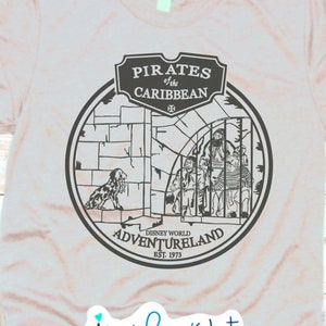 May include: A gray t-shirt with a black and white graphic of the Pirates of the Caribbean ride at Disney World's Adventureland. The graphic includes a pirate ship, a skull and crossbones, and the text "Pirates of the Caribbean" and "Adventureland".
