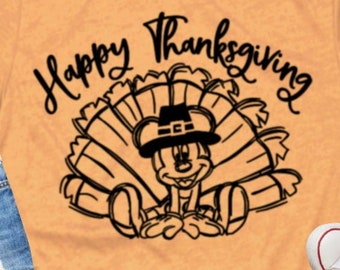 May include: Orange t-shirt with the text "Happy Thanksgiving" above a cartoon turkey character wearing a pilgrim hat. The shirt is paired with blue jeans and white sneakers.