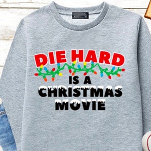 May include: Gray sweatshirt with the text "Die Hard is a Christmas Movie" in black letters with a string of colorful Christmas lights above the text.