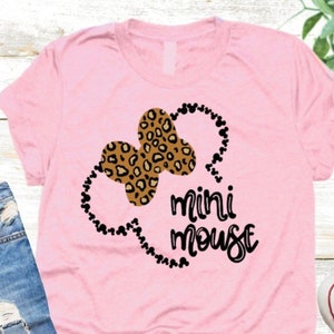 Puede incluir: Camiseta rosa con un contorno negro de las orejas de Mickey Mouse rellenas con un diseño de estampado de leopardo. El texto "mini mouse" está escrito en letras cursivas negras.