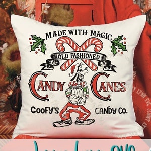 May include: A white pillow with a red and green Christmas design featuring a cartoon dog holding candy canes. The text reads "Made with Magic Old Fashioned Candy Canes Goofy's Candy Co."