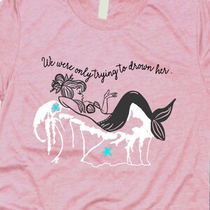 Puede incluir: Una camiseta rosa con una ilustración en blanco y negro de una sirena con el texto "We were only trying to drown her."
