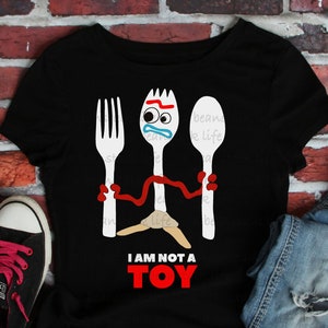 May include: Black t-shirt with a white graphic of a fork and spoon with faces. The text "I am not a toy" is printed below the graphic.