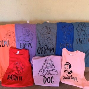 May include: Seven t-shirts in different colours and designs featuring the seven dwarfs from Snow White. The shirts have black text with the names of each dwarf: Sleepy, Happy, Grumpy, Dopey, Sneezy, Bashful, and Doc. One pink t-shirt features Snow White.