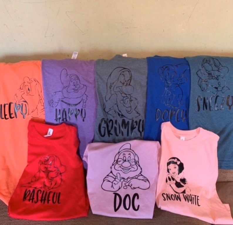 May include: Seven t-shirts in different colours and designs featuring the seven dwarfs from Snow White. The shirts have black text with the names of each dwarf: Sleepy, Happy, Grumpy, Dopey, Sneezy, Bashful, and Doc. One pink t-shirt features Snow White.