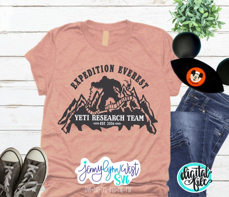 Pu&ograve; includere: Una maglietta rosa con un design grafico nero di una catena montuosa con uno yeti che cammina su un sentiero. Il testo "Expedition Everest Yeti Research Team - Est. 2006" &egrave; stampato sulla maglietta.