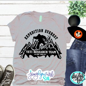 Pu&ograve; includere: Una maglietta grigia con un design grafico nero di uno yeti che scala una montagna con il testo "Expedition Everest Yeti Research Team Est. 2006"