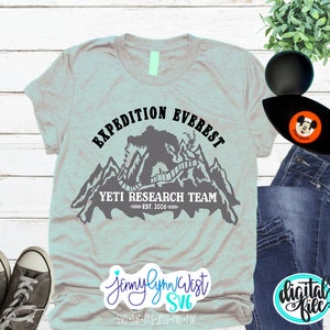 Pu&ograve; includere: Una maglietta grigia con un design grafico nero di uno Yeti in piedi di fronte a una catena montuosa. La grafica include il testo "EXPEDITION EVEREST", "YETI RESEARCH TEAM" e "EST. 2006".