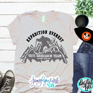 Pu&ograve; includere: Una maglietta grigia con un design grafico nero di uno Yeti in piedi su una catena montuosa. La grafica include il testo "Expedition Everest Yeti Research Team Est. 2006".