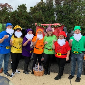 May include: Seven people dressed as the seven dwarfs from Snow White. They are wearing colourful shirts with their dwarf names on them. They are holding small cups and smiling.