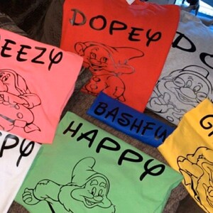 May include: Seven different coloured t-shirts with a black and white cartoon image of a dwarf and text on each shirt. The text on the shirts reads "Dopey", "Sneezy", "Bashful", "Happy", "Grumpy", "Doc", and "Sleepy".