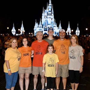 May include: A family of seven wearing matching t-shirts with the names of the seven dwarfs printed on them. The shirts are yellow, orange, red, and beige. The family are standing in front of Cinderella's Castle at Walt Disney World. The castle is lit up at night.