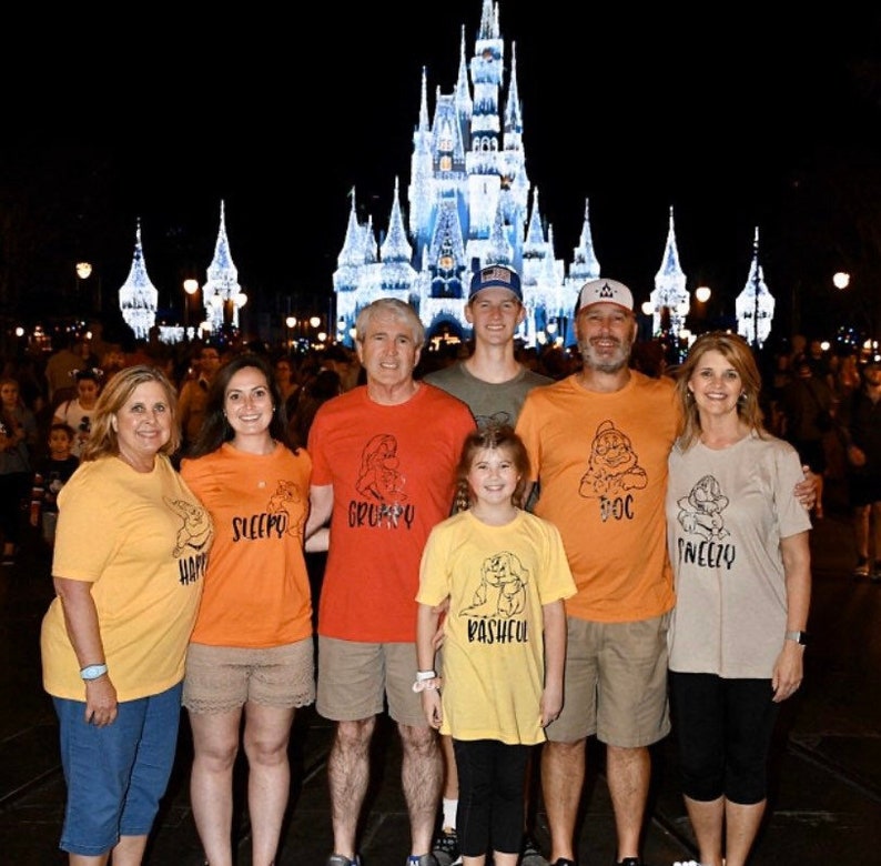 May include: A family of seven wearing matching t-shirts with the names of the seven dwarfs printed on them. The shirts are yellow, orange, red, and beige. The family are standing in front of Cinderella's Castle at Walt Disney World. The castle is lit up at night.
