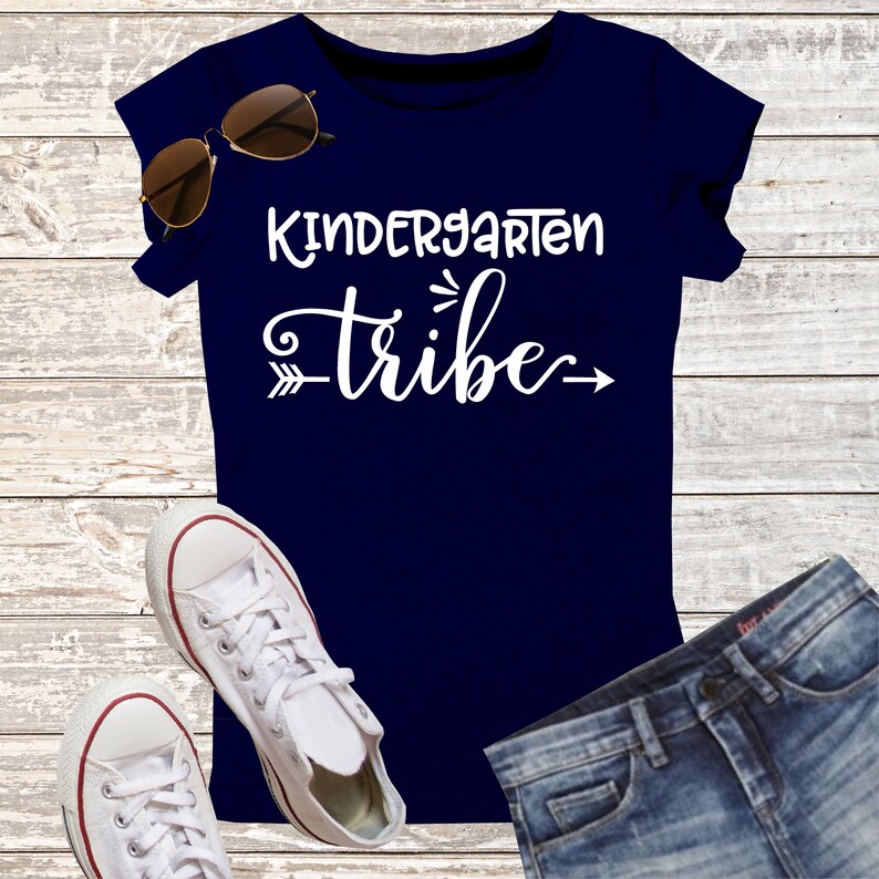 K&ouml;nnte beinhalten: Ein marineblaues T-Shirt mit dem wei&szlig;en Schriftzug "Kindergarten Tribe" und einem dekorativen Pfeil. Das Bild zeigt auch wei&szlig;e Turnschuhe, eine Sonnenbrille und Jeans, was auf ein Thema zum Schulanfang hindeutet.