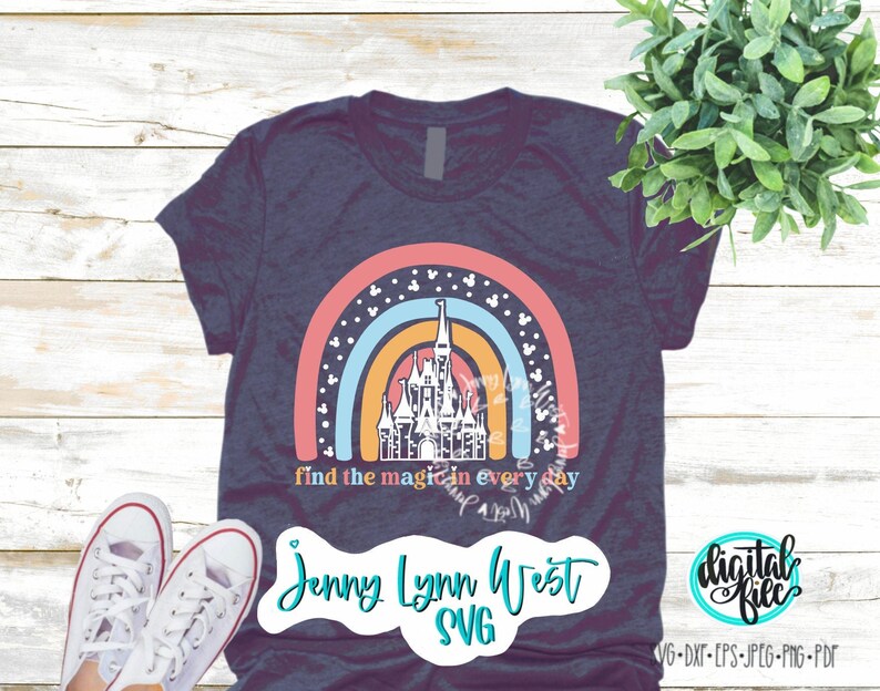 Op de afbeelding: Een marineblauw T-shirt met een regenboogontwerp met een wit silhouet van Assepoester's kasteel. De regenboog heeft witte stippen en de tekst "find the magic in every day". De onderkant van het shirt heeft de tekst "Jenny Lynn West SVG".