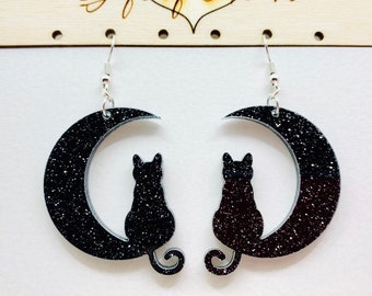 May include: A pair of black glitter cat earrings. Each earring features a black cat silhouette sitting on a crescent moon. The earrings hang from silver-colored hooks. The background is a light wooden sign with the text "Gift of Heart".