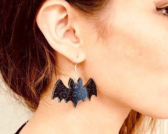 May include: A pair of bat-shaped earrings with a dark blue and glittery black design. The earrings are hanging from silver-colored hooks. The earrings are a Halloween-themed accessory.