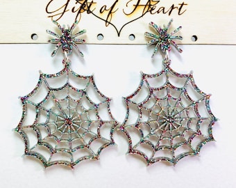 May include: A pair of silver-colored spiderweb earrings with iridescent glitter accents. Each earring features a spider at the top and a web design. The earrings are hanging against a white background.